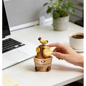 May include: A novelty wooden music box in the shape of a banana split. The music box has a brown wooden base with a smiling face, a yellow banana-shaped head, and a small banana on top. The word "Banana" is printed on the banana head.