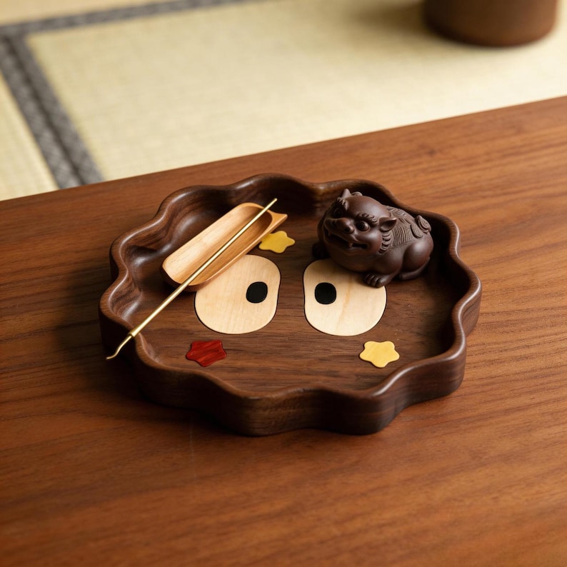 May include: A decorative wooden tray with a wavy edge, featuring a carved design. The tray holds a small brown figurine, a wooden scoop, and a gold-colored tool. The tray has inlaid wooden accents in red, yellow, and light brown.