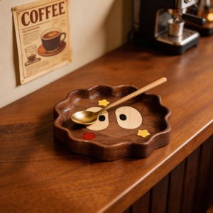 May include: A wooden, wavy-edged tray with a cartoon face design, featuring wooden eye and mouth accents, and yellow star details. A gold spoon rests on the tray. The tray is on a wooden surface, with a coffee-themed poster in the background.