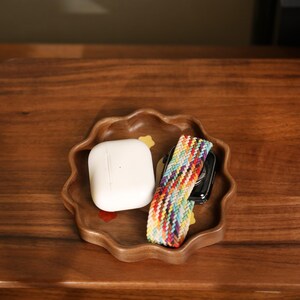 May include: A wooden, scalloped-edge tray holds a white wireless earbud case, a colorful woven watch band, and a black smartwatch. The tray is brown and sits on a wooden surface.