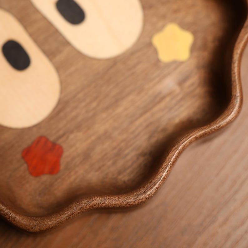 May include: A wooden serving tray with a dark brown finish. The tray features a wavy edge and is decorated with light-colored wooden shapes and red and yellow star-shaped inlays. The tray is on a wooden surface.