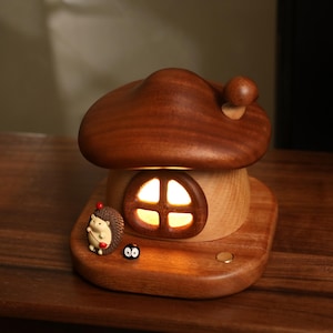 May include: A wooden mushroom-shaped night light with a warm glow emanating from the window. The lamp features a brown cap and a light-colored base. A small hedgehog figurine and a black soot sprite are placed on the base.