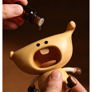 May include: A wooden figurine with a surprised expression, mouth open, and a single wooden tooth. A small brown glass bottle is held above the figurine, with a drop of liquid falling towards the open mouth. The figurine has black dot eyes and small arms.