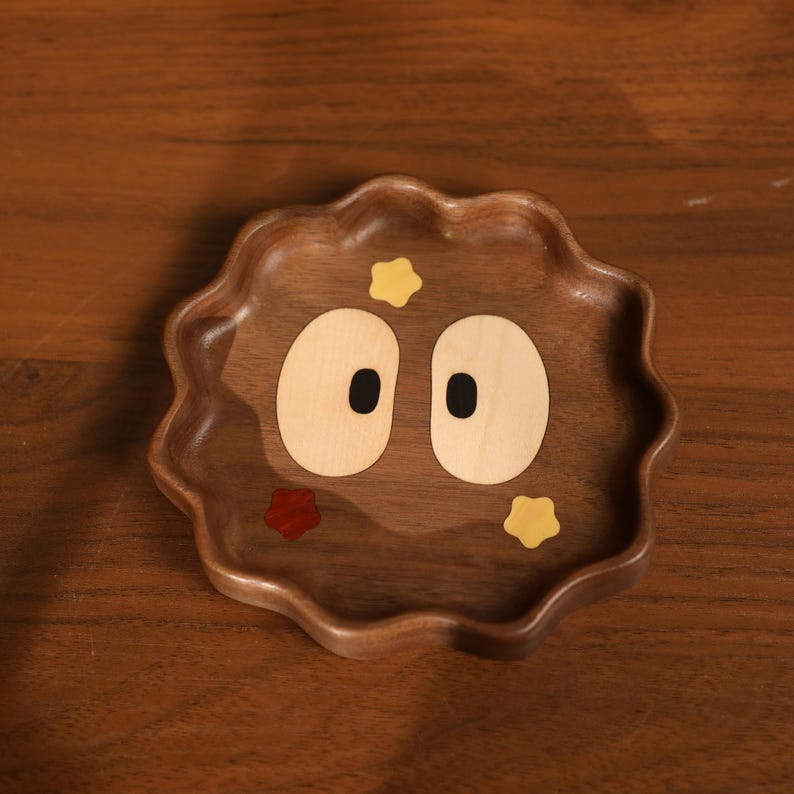 May include: A wooden tray with a whimsical face design. The tray is a dark brown color with a wavy edge. The face features two large, oval-shaped eyes, and small star and red accents.