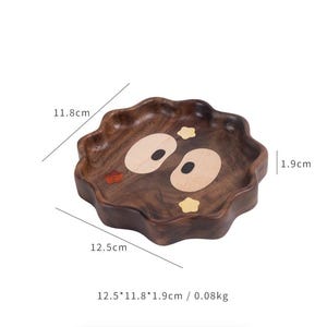 May include: A wooden tray with a wavy edge and a cartoon face design. The tray is brown with light-colored oval eyes and small yellow accents. It measures 12.5 cm x 11.8 cm x 1.9 cm.