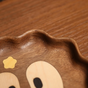 May include: A wooden serving tray with a bear face design. The tray is made of dark brown wood and features a light-colored star and bear face details. The edges of the tray have a wavy, scalloped design.