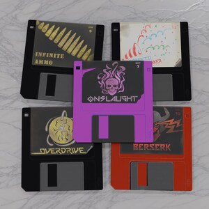 Könnte beinhalten: Fünf Retro-Disketten mit verschiedenen Designs. Eine Diskette ist lila mit einem Totenkopf und dem Wort "ONSLAUGHT". Andere Disketten zeigen Texte wie "INFINITE AMMO" und "OVERDRIVE". Die Disketten sind schwarz, rot und lila.