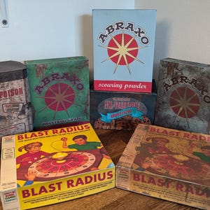 May include: A collection of vintage-style boxes and tins. The items include boxes labeled "ABRAXO" and "BLAST RADIUS", and a tin marked "RAT-POISON". The boxes feature retro graphics and text, with a weathered, aged appearance.
