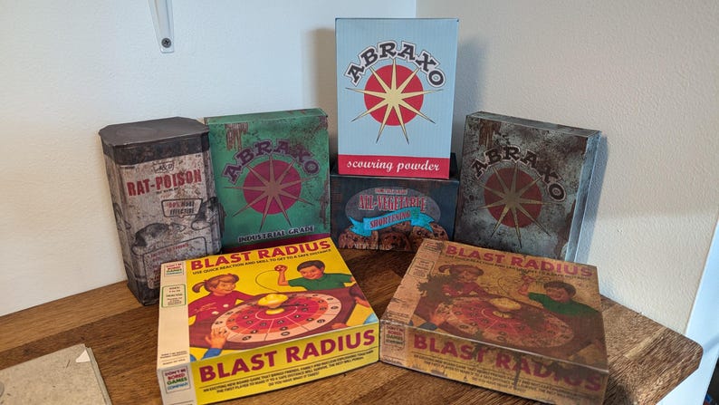 May include: A collection of vintage-style boxes and tins. The items include boxes labeled "ABRAXO" and "BLAST RADIUS", and a tin marked "RAT-POISON". The boxes feature retro graphics and text, with a weathered, aged appearance.