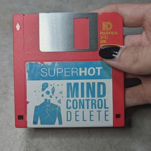 Op de afbeelding: Een rode 3,5-inch diskette met een zilveren schuifbare metalen hoes. Het label luidt "SUPERHOT MIND CONTROL DELETE" met een blauwe afbeelding van een persoon. De diskette wordt door een hand vastgehouden.
