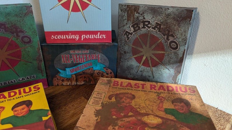 May include: A collection of vintage-style boxes with distressed textures and retro graphics. The boxes feature text like "scouring powder," "All-Vegetarite Shortening," and "Blast Radius." The color palette includes red, green, and yellow.