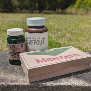 Peut inclure: Trois articles sur le thème de Fallout : une bouteille en verre vert étiquetée "RAD-X", une bouteille marron marquée "ORIGINAL BUFFOUT" et une boîte en carton avec "MENTATS" imprimé dessus. Les articles sont posés sur une surface en pierre avec un arrière-plan vert flou.