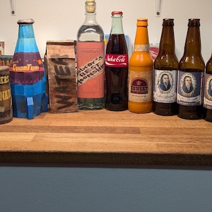 Puede incluir: Una colección de botellas y latas de estilo vintage, que incluye una botella azul y roja, una botella de Coca-Cola y varias botellas de cerveza marrones con etiquetas de retrato. Los artículos están dispuestos sobre una superficie de madera.