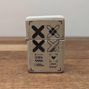May include: A white, rectangular lighter with black graphic designs. The top half features two black "X" shapes and a square pattern. The bottom half has more symbols, including a heart and text that reads "THANKFUL WITH DARE".