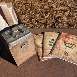 Pode incluir: Uma mala de couro castanho desgastado com uma pega de metal e uma tampa articulada, contendo um dispositivo de metal com botões e mostradores. Ao lado, três livretos envelhecidos, um intitulado "Who Goes There?" com texto.