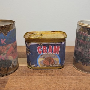 Può includere: Tre barattoli di cibo vintage con etichette danneggiate. I barattoli sono etichettati "Pork N Beans", "CRAM" e "Dog Shoes". I barattoli hanno un aspetto arrugginito e invecchiato.