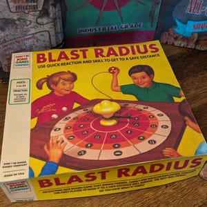 May include: A vintage board game box titled "BLAST RADIUS" with illustrations of children playing. The box is yellow with red and brown accents. Other boxes are visible in the background, suggesting a collection of vintage games.