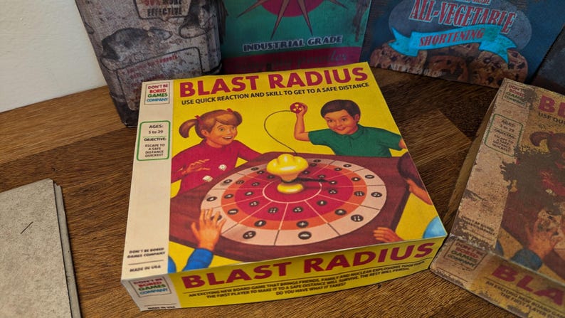 May include: A vintage board game box titled "BLAST RADIUS" with illustrations of children playing. The box is yellow with red and brown accents. Other boxes are visible in the background, suggesting a collection of vintage games.