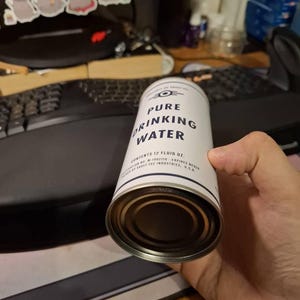 Puede incluir: Una lata blanca y plateada de "Pure Drinking Water" con texto negro. La lata tiene una banda azul y blanca en la parte superior e inferior. La lata contiene 355 ml de agua.