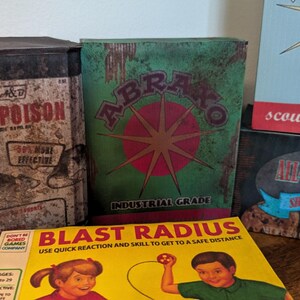 May include: A collection of vintage-style boxes and signs. The items include a Rat-Poison container, a green box with "ABRAKO" and a starburst design, and a yellow box with "BLAST RADIUS" text and illustrations. Also, a sign that says "scouring powder".