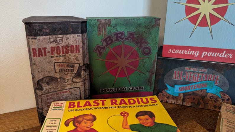 May include: A collection of vintage-style boxes and signs. The items include a Rat-Poison container, a green box with "ABRAKO" and a starburst design, and a yellow box with "BLAST RADIUS" text and illustrations. Also, a sign that says "scouring powder".