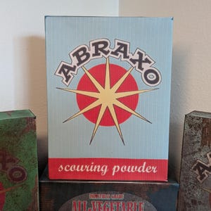 May include: Three vintage-style boxes with the word "ABRAXO" in a retro font. The central box is light blue with a red and yellow starburst design and the words "scouring powder". The other boxes are green and brown.