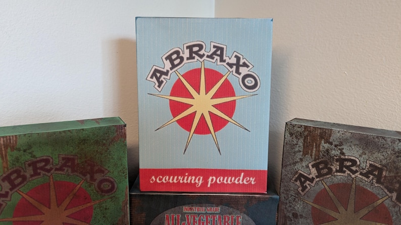 May include: Three vintage-style boxes with the word "ABRAXO" in a retro font. The central box is light blue with a red and yellow starburst design and the words "scouring powder". The other boxes are green and brown.