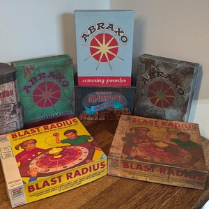 May include: A collection of vintage-style boxes and a tin. The boxes feature the text "ABRAXO" and "BLAST RADIUS" with colorful graphics. One tin reads "RAT-POISON". The items are arranged on a wooden surface.