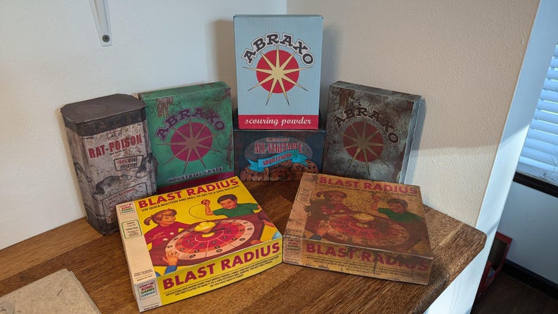 May include: A collection of vintage-style boxes and a tin. The boxes feature the text "ABRAXO" and "BLAST RADIUS" with colorful graphics. One tin reads "RAT-POISON". The items are arranged on a wooden surface.