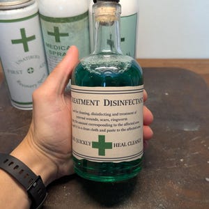 May include: A vintage-style glass bottle filled with green liquid, labeled "Treatment Disinfectant." The label includes instructions and a green cross symbol. Other medical spray bottles are in the background.