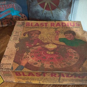 May include: A vintage-style board game box titled "Blast Radius" with illustrations of children playing. The box is brown with red and yellow accents. Other boxes with similar themes are visible in the background.