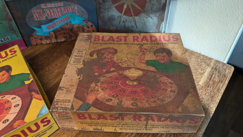 May include: A vintage-style board game box titled "Blast Radius" with illustrations of children playing. The box is brown with red and yellow accents. Other boxes with similar themes are visible in the background.