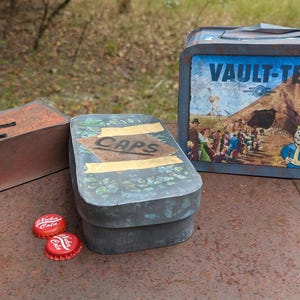 May include: A selection of Fallout-themed items, including a rectangular box, a metal container labeled "CAPS", a lunchbox with "VAULT-TEC" text, and two red bottle caps. The items are arranged on a rusty metal surface.
