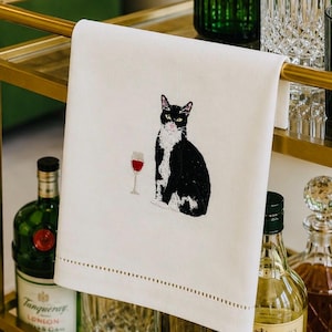 May include: A white rectangular kitchen towel featuring an embroidered black and white tuxedo cat sitting next to a glass of red wine. The towel has a decorative hemstitch detail along the bottom edge and is draped over a gold metal bar.