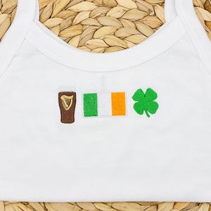 May include: White tank top with embroidered symbols: a brown pint of beer, the Irish flag, and a green four-leaf clover. The tank top has a scoop neck and thin straps.