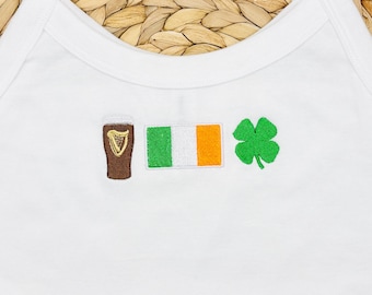 May include: A white tank top featuring three embroidered designs centered on the chest: a brown pint glass with a gold harp, the green, white, and orange Irish flag, and a bright green four-leaf clover. The garment has a round neckline.