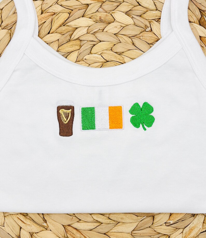 May include: White tank top with embroidered symbols: a brown pint of beer, the Irish flag, and a green four-leaf clover. The tank top has a scoop neck and thin straps.