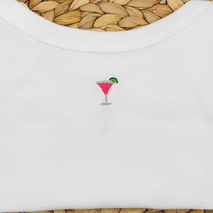 May include: White t-shirt featuring an embroidered cocktail design. The cocktail glass is pink with a green lime wedge. The shirt is made of a soft, white fabric.