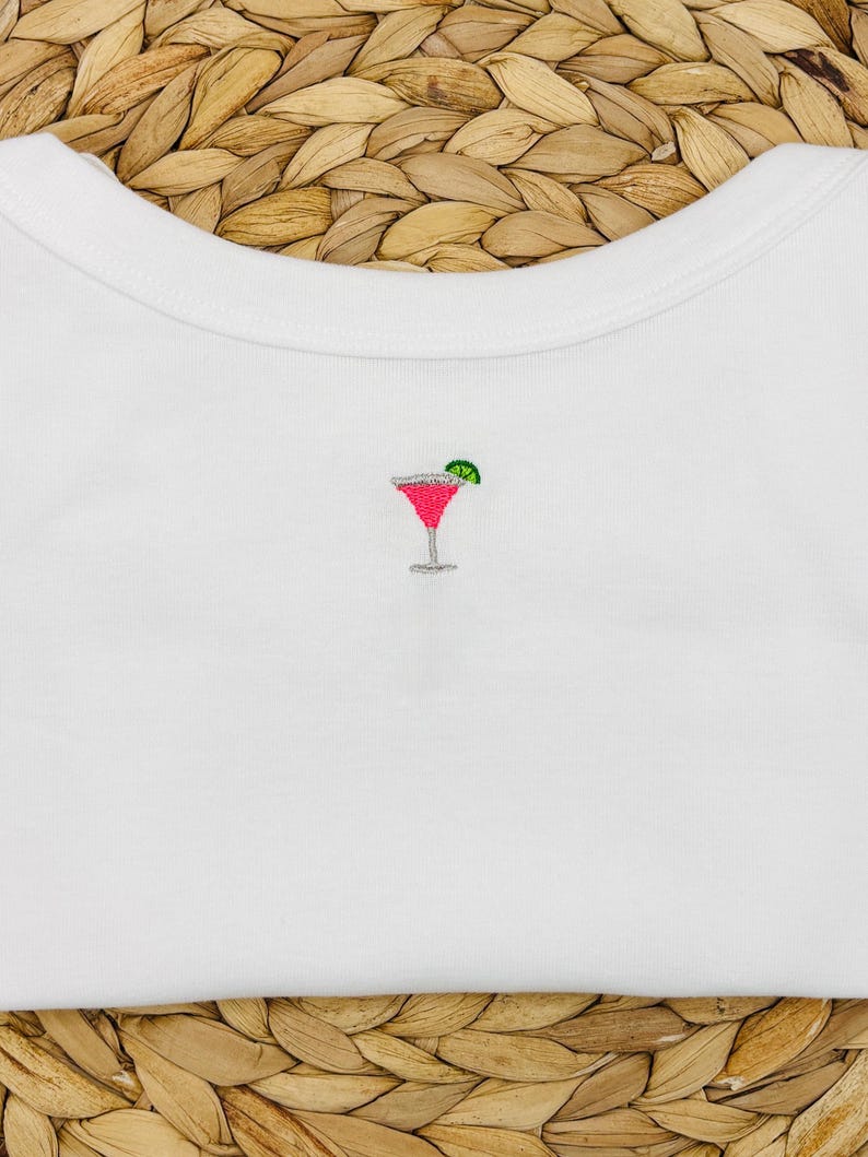 May include: White t-shirt featuring an embroidered cocktail design. The cocktail glass is pink with a green lime wedge. The shirt is made of a soft, white fabric.