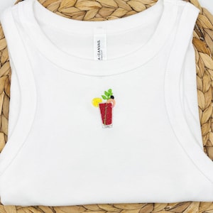 May include: White vest top featuring an embroidered cocktail design. The cocktail is red with a lemon slice and green garnish. The vest is sleeveless and made of a soft material. The tag reads "A. CANVAS."