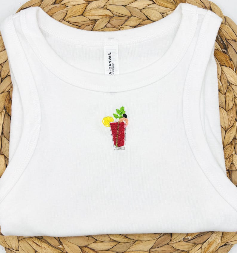 May include: White vest top featuring an embroidered cocktail design. The cocktail is red with a lemon slice and green garnish. The vest is sleeveless and made of a soft material. The tag reads "A. CANVAS."