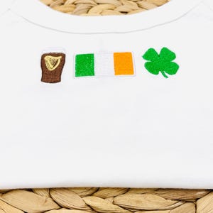 May include: White baby bodysuit with embroidered St. Patrick's Day themed designs. Features a brown pint of stout, the Irish flag, and a green shamrock. The bodysuit is made of soft, white fabric.