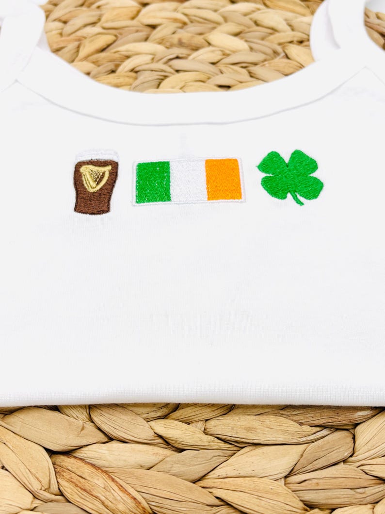 May include: White baby bodysuit with embroidered St. Patrick's Day themed designs. Features a brown pint of stout, the Irish flag, and a green shamrock. The bodysuit is made of soft, white fabric.