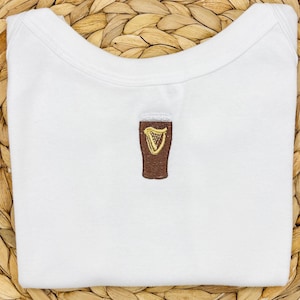 May include: White baby onesie with a brown embroidered pint of beer and a gold harp design. The onesie is folded neatly. The beer glass is centered on the front of the garment. The onesie is made of soft material.