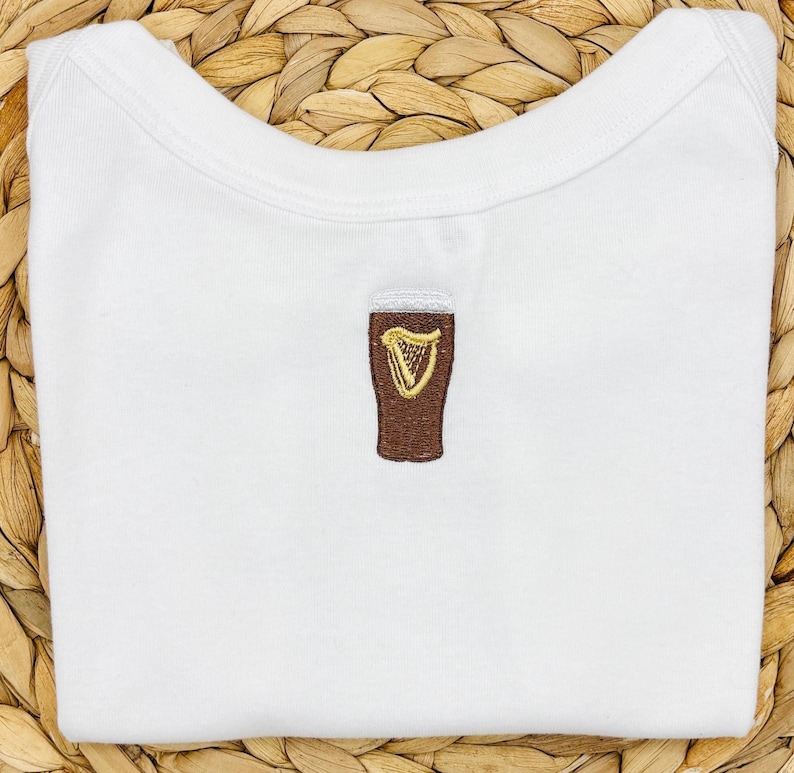 May include: White baby onesie with a brown embroidered pint of beer and a gold harp design. The onesie is folded neatly. The beer glass is centered on the front of the garment. The onesie is made of soft material.