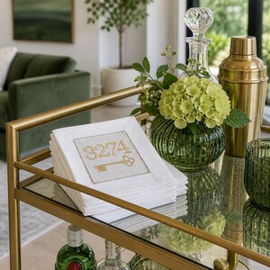May include: A gold bar cart with a glass top displays a stack of white napkins embroidered with the number "3274" and a key design in gold thread. A green floral arrangement, a gold cocktail shaker, and glassware complete the elegant setup.