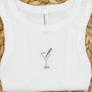 May include: White vest top featuring a small embroidered martini glass design. The martini glass has a silver outline, a black olive, and a toothpick. The vest top is made of a soft material.