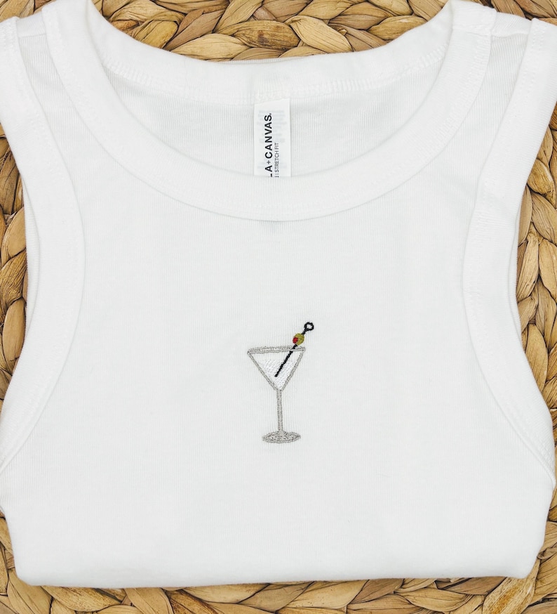 May include: White vest top featuring a small embroidered martini glass design. The martini glass has a silver outline, a black olive, and a toothpick. The vest top is made of a soft material.
