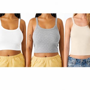 May include: Three cropped tank tops in white, grey, and beige. The white tank top has spaghetti straps and a scoop neckline. The grey tank top has a racerback design. The beige tank top has thin straps and a scoop neckline. All three tops are made of a ribbed material.