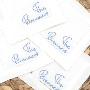 May include: Four square white fabric napkins featuring a hemstitched border and the text "The Brenners" embroidered in light blue cursive script. Each napkin is identical in design, showcasing a clean, white textile with blue thread.